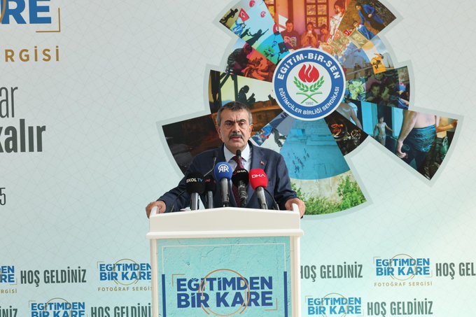 Yusuf Tekin stands at a podium with multiple microphones, delivering a speech. A large banner behind him reads "Eğitimden Bir Kare Fotoğraf Sergisi" and displays photos of educational activities. A crowd of people is gathered, listening attentively. The setting appears to be an outdoor area at Ankara ATG YHT Gar.