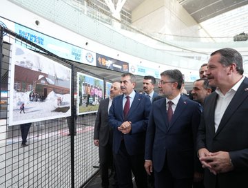 Yusuf Tekin, İbrahim Yumaklı, and Ali Yalçın in suits with red ties, standing in a modern indoor space with large photo displays. The photos show children in educational settings and damaged buildings, mounted on metal frames. Banners display "Eğitimden Bir Kare Fotoğraf Sergisi" and images of people in various activities.