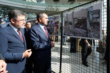 Yusuf Tekin, İbrahim Yumaklı, and Ali Yalçın in suits with red ties, standing in a modern indoor space with large photo displays. The photos show children in educational settings and damaged buildings, mounted on metal frames. Banners display "Eğitimden Bir Kare Fotoğraf Sergisi" and images of people in various activities.