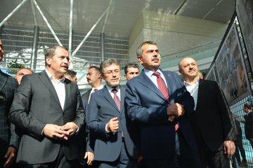 Yusuf Tekin, İbrahim Yumaklı, and Ali Yalçın in suits with red ties, standing in a modern indoor space with large photo displays. The photos show children in educational settings and damaged buildings, mounted on metal frames. Banners display "Eğitimden Bir Kare Fotoğraf Sergisi" and images of people in various activities.