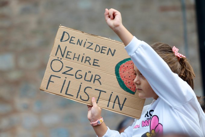 A child holding a cardboard sign with handwritten text in Turkish reading "DENIZDEN NEHIRE OZGU FILISTIN" and a drawing of a watermelon. The child is wearing a white sweatshirt and has a hair bow, raising one fist.