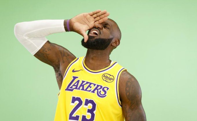 LeBron James con camiseta amarilla de Los Angeles Lakers con el número 23, manga blanca en el brazo izquierdo, muñequera morada, tatuajes en el brazo derecho, barba corta, mano levantada a la cara en gesto de celebración, fondo verde.