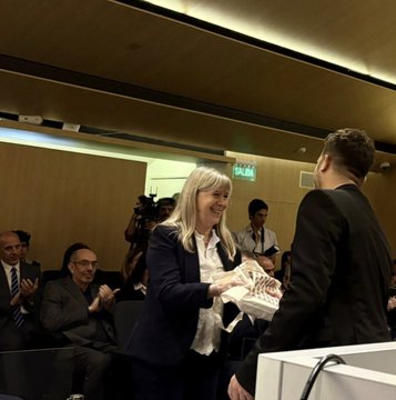 First image shows a woman in beige pants and blouse holding a tablet standing near a podium in a modern courtroom with wooden walls and Argentine flag behind banners for Programa de Justicia Sustentable and CMM logo. Second image depicts a bald man in blue suit handing a document to a younger man in blue shirt with another man clapping seated nearby and a woman in purple top at the bench area with similar banners and flag. Third image features a silver-haired woman in navy suit handing a fabric-wrapped item to a young man in black suit amid audience members including men in suits and a photographer with exit sign visible on wooden paneled room.