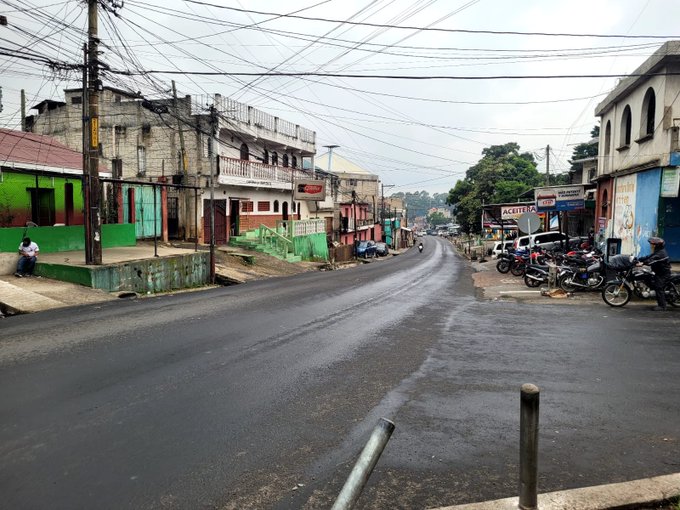 A wet street in an urban neighborhood lined with colorful buildings in shades of green blue red and yellow some with graffiti and advertisements parked motorcycles cars and a white vehicle visible along the roadside power lines overhead and trees in the background under overcast skies