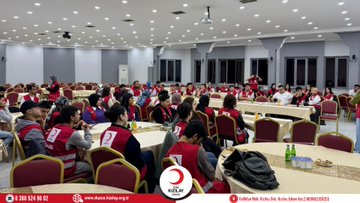 First image shows a conference room with participants seated around tables wearing red vests some standing and one woman presenting in front of a projector screen displaying Turkiye Afet Müdahale Ekip with Turkish Red Crescent logo visible on screen and tables. Second image depicts a larger group of people in red vests seated at round tables in a spacious hall with beverages on tables and the Turkish Red Crescent logo on a banner. Third image features a group photo of diverse participants including men and women in red vests standing together in a room with the Turkish Red Crescent logo on a banner behind them. Watermarks from Düzce Kızılay appear on all images.