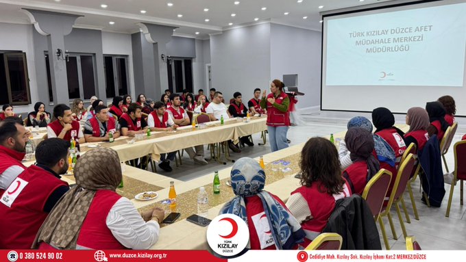 First image shows a conference room with participants seated around tables wearing red vests some standing and one woman presenting in front of a projector screen displaying Turkiye Afet Müdahale Ekip with Turkish Red Crescent logo visible on screen and tables. Second image depicts a larger group of people in red vests seated at round tables in a spacious hall with beverages on tables and the Turkish Red Crescent logo on a banner. Third image features a group photo of diverse participants including men and women in red vests standing together in a room with the Turkish Red Crescent logo on a banner behind them. Watermarks from Düzce Kızılay appear on all images.