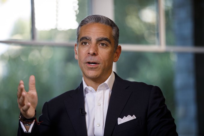 Man with gray hair wearing a dark suit white shirt and pocket square speaks into a microphone in a bright room with large windows showing greenery outside. Second image shows white Bitcoin symbol on orange background.