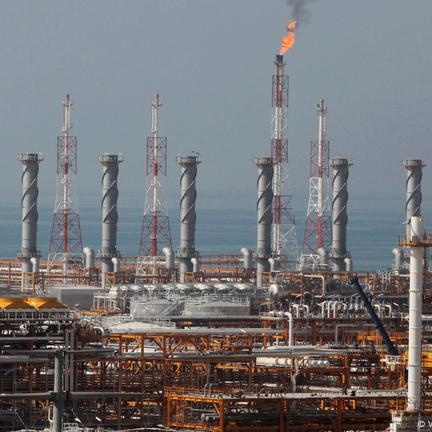An industrial facility with multiple tall cylindrical structures and pipes, featuring a gas flare emitting a flame into the sky. The facility is situated near a body of water, with a complex network of metal frameworks and equipment visible. The Iranian flag, displaying green, white, and red horizontal stripes with a red emblem and stylized text, is shown above the facility.
