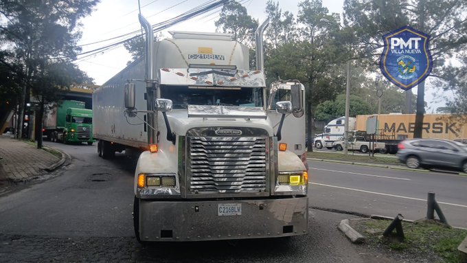 A white semi-trailer truck with mechanical failure is parked on a multi-lane road at the entrance to Anillo Periferico, obstructing northbound traffic in Aguilar Batres. The truck displays Despacho logo on its side and has yellow lights on. Surrounding vehicles include green and white trucks, a silver car, and a bus. A blue sign reading PMT is visible nearby, indicating local transit police presence. Trees and a sidewalk line the roadside.