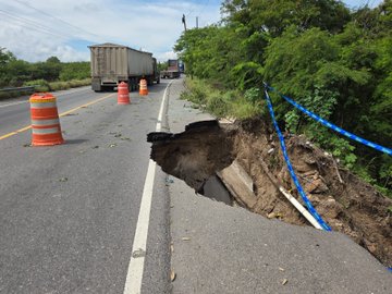 First image shows a large truck parked on a two-lane highway with orange traffic cones placed around a significant subsidence in the asphalt, exposing dirt and rocks below, surrounded by green bushes and trees under a clear sky. Second image depicts a close-up of the deep subsidence with jagged asphalt edges, blue caution tape draped around the hole, scattered debris including broken concrete slabs and grass on the roadside. Third image captures the subsidence from a wider angle with yellow and blue caution tapes stretched across the damaged section of the road, green fields and trees in the background, and a road sign visible nearby. Fourth image illustrates the subsidence area with blue caution tape, a truck in the distance on the highway, and surrounding vegetation including bushes and trees.