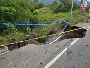 First image shows a large truck parked on a two-lane highway with orange traffic cones placed around a significant subsidence in the asphalt, exposing dirt and rocks below, surrounded by green bushes and trees under a clear sky. Second image depicts a close-up of the deep subsidence with jagged asphalt edges, blue caution tape draped around the hole, scattered debris including broken concrete slabs and grass on the roadside. Third image captures the subsidence from a wider angle with yellow and blue caution tapes stretched across the damaged section of the road, green fields and trees in the background, and a road sign visible nearby. Fourth image illustrates the subsidence area with blue caution tape, a truck in the distance on the highway, and surrounding vegetation including bushes and trees.