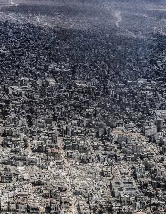 Aerial view from high altitude showing extensive urban destruction in a densely built area with numerous collapsed and damaged buildings spread across a wide landscape under a clear sky with a river or waterway visible in the background amid the rubble and debris