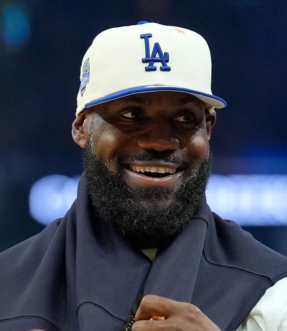 Un hombre con barba que lleva una gorra blanca de los Dodgers de Los Ángeles con letras azules de Los Ángeles y un parche de campeonato sonríe mientras está vestido con una camisa azul oscuro y pantalones claros en un escenario de arena cubierta con poca luz.