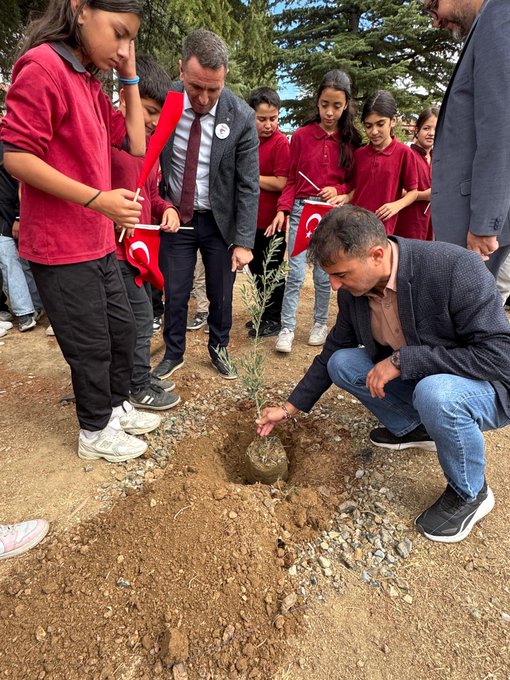First image shows exterior of a school building with yellow and blue walls, sign reading Bağıllı Şehit Feyyaz Korkmaz Ortaokulu, multiple Turkish flags and Palestinian flags held by hands in foreground against blue sky. Second image depicts a man in gray suit and tie sitting at table with two girls in school uniforms, table covered with colorful bracelets in red white black green resembling Palestinian flag colors, two Palestinian flags draped on table edges, trees and motorcycle in background. Third image captures group of children in red uniforms and adults in suits around a small hole in dirt where a man in beige shirt kneels planting a sapling, Turkish flags held by children, olive branches nearby, dirt ground with trees in background. Fourth image features large group of children mostly in red uniforms and adults standing on dirt field, many holding red Turkish flags, cloudy sky above with trees and yellow building in background.