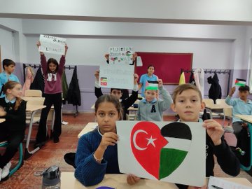 First image shows a group of schoolchildren in a classroom holding up handmade signs with messages like YURTTA BARIS DUNYADA BARIS and Palestinian flags some wearing keffiyehs standing near desks with books and bags. Second image depicts children hands folding and displaying paper Palestinian flags on a table with crayons and blue paper sheets scattered around. Third image captures a television screen mounted on a wall in a school hallway broadcasting a waving Palestinian flag against a cloudy sky with decorative plants and bulletin boards nearby. Fourth image features schoolchildren in another classroom holding signs reading FILISTIN and combined Turkish-Palestinian flags some wearing headscarves standing by desks and a chalkboard.