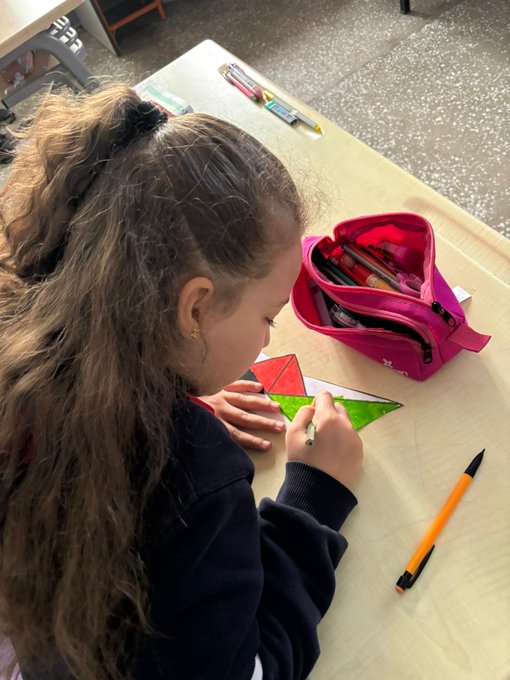 First image shows a group of about 20 uniformed school children standing in a classroom holding up large handmade Palestinian flags some children wear face masks others stand attentively near wooden desks and cabinets with a bulletin board in the background. Second image depicts a young girl with long brown hair in a ponytail wearing a school uniform sitting at a wooden table drawing on green and red paper shapes using colored pencils and markers from a pink pencil case nearby. Third image captures a large group of black-uniformed students arranged in the shape of a heart on a red-paved school courtyard surrounded by buildings trees and mountains under a cloudy sky with a Turkish flag visible. Fourth image features a black bulletin board decorated with white peace doves multiple colorful handmade Palestinian flags and cutout shapes with the Turkish text ÖZGÜRLÜĞÜN FİLİSTİNİN written in large letters across the top.