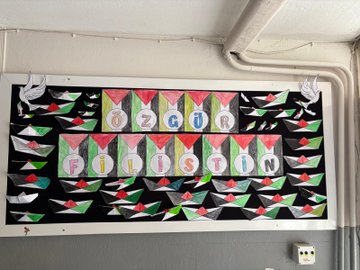 First image shows a group of about 20 uniformed school children standing in a classroom holding up large handmade Palestinian flags some children wear face masks others stand attentively near wooden desks and cabinets with a bulletin board in the background. Second image depicts a young girl with long brown hair in a ponytail wearing a school uniform sitting at a wooden table drawing on green and red paper shapes using colored pencils and markers from a pink pencil case nearby. Third image captures a large group of black-uniformed students arranged in the shape of a heart on a red-paved school courtyard surrounded by buildings trees and mountains under a cloudy sky with a Turkish flag visible. Fourth image features a black bulletin board decorated with white peace doves multiple colorful handmade Palestinian flags and cutout shapes with the Turkish text ÖZGÜRLÜĞÜN FİLİSTİNİN written in large letters across the top.