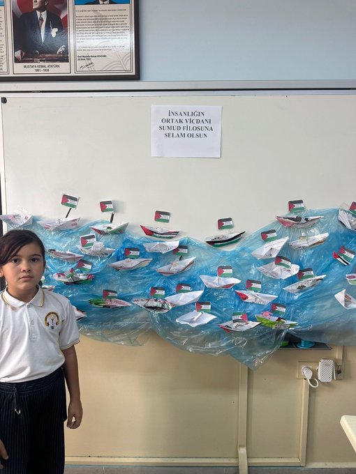 First image shows a classroom wall display with numerous small paper boats in blue and white, adorned with UAE flags, arranged in rows against a blackboard with Turkish text reading SUMINSI OLANA SELAM OLANA and a Turkish flag. Second image depicts a framed portrait of Mustafa Kemal Ataturk on the wall, a sign in Turkish saying INSANLIK SUMINSI SELAM SUMINSI, and a girl in school uniform standing beside a blue wavy display of paper boats with UAE flags. Third image captures two girls in white uniforms sitting at a table drawing on large paper sheets depicting a colorful world map with peace doves, Palestinian and other flags, butterflies, and green plants using colored pencils. Fourth image features a classroom wall with a sign in Turkish INSAN OLANA SUM OLANA SELAM OLANA, a girl in uniform pointing to a blue wavy display of paper boats decorated with UAE flags.