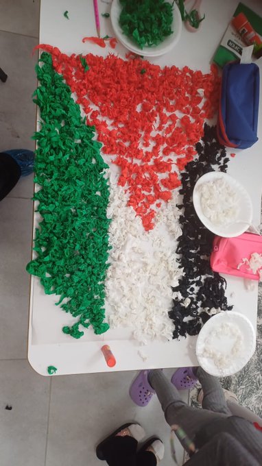 First image shows a bulletin board with colorful handprints in red green black and white forming Palestinian flag patterns around a central green watermelon illustration and text in Turkish reading Nehirden nehirden başlar Filistin with paper butterflies in flag colors attached. Second image depicts a table with shredded colored paper in red black white and green arranged to form the Palestinian flag shape bowls of paper scraps glue sticks and childrens feet visible indicating craft activity. Third image features a black background covered in paper butterflies folded from colored paper in red white green and black matching flag colors with drawn hands in pink holding them open. Fourth image captures children in a classroom sitting at yellow tables holding paper plate crafts resembling boats or masks painted in blue with decorations wearing head coverings in Palestinian flag colors surrounded by classroom shelves and supplies.