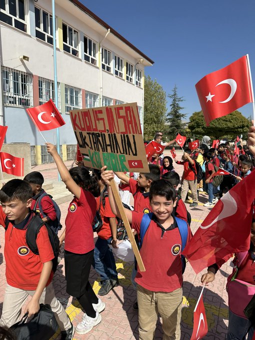 First image shows a hand-drawn artwork on paper depicting a figure in black niqab with long flowing hair, eyes visible, wearing a red shoulder piece, surrounded by green areas resembling the Palestinian map outline, with two heart stickers one Turkish flag and one Palestinian flag attached nearby. Second image displays a young boy in a wheelchair inside a school room with orange walls and shelves of bags, holding a Palestinian flag with black white green red stripes, dressed in a checkered scarf and green outfit. Third image captures a smiling girl with face painted in black white green red stripes on cheeks, holding a Palestinian flag, standing on stairs with other children some holding signs reading Hepimiz Gazze in Turkish. Fourth image portrays a group of school children in red uniforms marching outdoors near a building, waving Turkish flags, one child holding a sign reading Ozgurlukte Insanlik Var in Turkish.