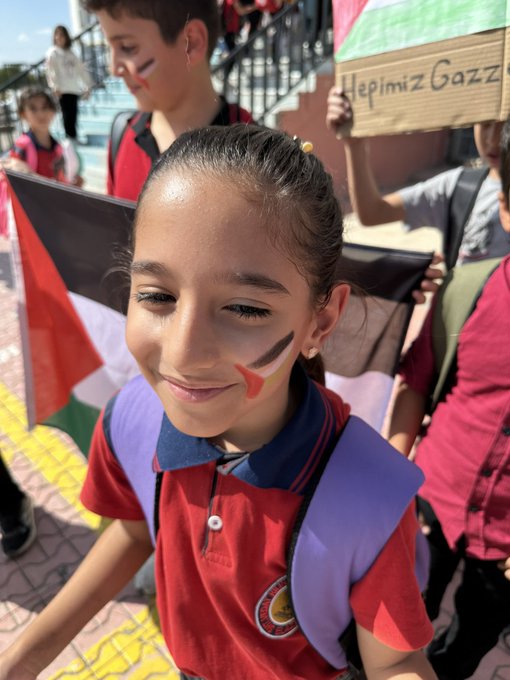 First image shows a hand-drawn artwork on paper depicting a figure in black niqab with long flowing hair, eyes visible, wearing a red shoulder piece, surrounded by green areas resembling the Palestinian map outline, with two heart stickers one Turkish flag and one Palestinian flag attached nearby. Second image displays a young boy in a wheelchair inside a school room with orange walls and shelves of bags, holding a Palestinian flag with black white green red stripes, dressed in a checkered scarf and green outfit. Third image captures a smiling girl with face painted in black white green red stripes on cheeks, holding a Palestinian flag, standing on stairs with other children some holding signs reading Hepimiz Gazze in Turkish. Fourth image portrays a group of school children in red uniforms marching outdoors near a building, waving Turkish flags, one child holding a sign reading Ozgurlukte Insanlik Var in Turkish.