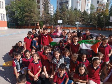 Children in school uniforms form the number 100 using their bodies on a colorful playground with basketball hoop and surrounding buildings and trees. A group of children in uniforms hold a Palestinian flag under trees with a man in white shirt nearby. A crowd of children in red uniforms and a woman in hijab pose smiling with Palestinian flags on the schoolyard near a basketball hoop. A bulletin board displays childrens crayon drawings of ships with Palestinian and Turkish flags, the word Gazze, and multicolored solidarity messages.