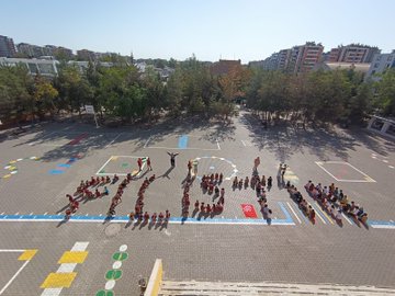 Children in school uniforms form the number 100 using their bodies on a colorful playground with basketball hoop and surrounding buildings and trees. A group of children in uniforms hold a Palestinian flag under trees with a man in white shirt nearby. A crowd of children in red uniforms and a woman in hijab pose smiling with Palestinian flags on the schoolyard near a basketball hoop. A bulletin board displays childrens crayon drawings of ships with Palestinian and Turkish flags, the word Gazze, and multicolored solidarity messages.