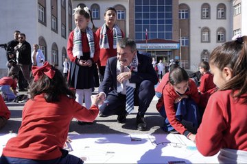 First image shows a man in a suit kneeling on the ground in a school courtyard, shaking hands with young girls in red uniforms and headscarves while other children draw on large paper sheets depicting Palestinian flags and symbols. Second image depicts a group of suited men and children in uniforms standing before several easels displaying paintings including a woman in a headscarf resembling Palestinian figures and abstract art with breaking chains and raised fists. Third image captures a classroom scene with a man in a suit speaking near a projector screen, a woman in white coat assisting, and girls in uniforms seated at desks attentively watching. Fourth image features a large group of adults in professional attire including white coats and suits posing in front of a school building with a Turkish flag and sign reading Ömer Duygun İlkokulu.