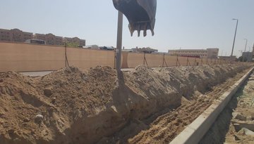 First image depicts a rendered design of a purple walking path lined with artistic floral and fish motifs, covered by white arched canopies, adjacent to a road with buildings and sand in the background under a sunset sky. Second image shows construction site with an excavator bucket extended over a trench filled with sand, surrounded by fences, buildings, and streetlights in a sunny urban area. Third image illustrates an ongoing construction area with multiple buildings including a green Top Green structure, excavator, sand piles, fences, and debris under a clear blue sky.
