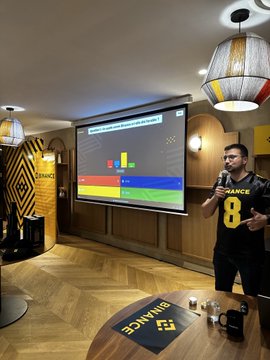First image shows a man in a black and yellow Binance jersey numbered 8 standing in front of a projector screen displaying a quiz interface with colored bars and graphs in a modern room with wooden floors yellow walls pendant lamps and Binance logos on the wall and table. Second image depicts the same man from behind facing an audience of diverse attendees seated in a large room with wooden flooring and lighting fixtures engaging in the event. Third image captures a stage setup with a trophy podium and screen showing Le Grand Quiz Crypto by Binance with an audience seated in chairs under a glass ceiling in a spacious venue with plants and modern decor.