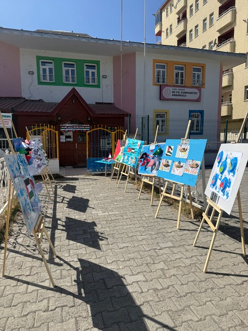 Multiple images show outdoor school courtyard with colorful buildings in pastel shades including pink green and yellow facades with Turkish flags. Easels display childrens vibrant paintings of flags animals and abstract designs in blues reds and greens. Tables hold folded paper boats in Turkish and Palestinian flag colors with some featuring Sumud Filosu labels. Palestinian flags are visible on boards and displays alongside educational posters.