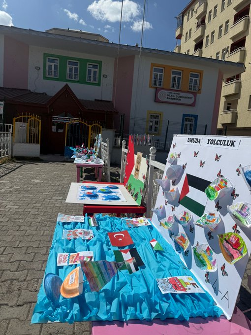 Multiple images show outdoor school courtyard with colorful buildings in pastel shades including pink green and yellow facades with Turkish flags. Easels display childrens vibrant paintings of flags animals and abstract designs in blues reds and greens. Tables hold folded paper boats in Turkish and Palestinian flag colors with some featuring Sumud Filosu labels. Palestinian flags are visible on boards and displays alongside educational posters.