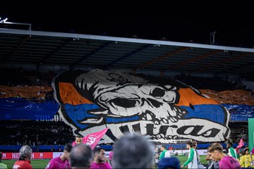 Large fabric banner with white skull and orange outlines displayed in dark stadium stands surrounded by blue and orange fabric sections fans in pink shirts visible below and to the sides green field and advertising boards in background. Long horizontal banner with French text Vous navez peur jamais sans la haine in white letters on blue background stretched across stands with red smoke and flares from fans below goalpost structure and sponsor ads on pitch side. Silhouetted figures of fans in hoodies raising arms holding lit red flares creating bright red glow and thick red smoke against metal railing in dimly lit stadium section. Fabric banner with bold white text Jamais sans la haine draped over stands near goal net with dark fabric sections and smoke wisps players in green jerseys on green field below sponsor logos like TNT and BKT visible.