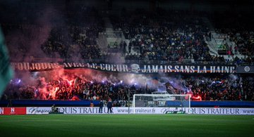 Large fabric banner with white skull and orange outlines displayed in dark stadium stands surrounded by blue and orange fabric sections fans in pink shirts visible below and to the sides green field and advertising boards in background. Long horizontal banner with French text Vous navez peur jamais sans la haine in white letters on blue background stretched across stands with red smoke and flares from fans below goalpost structure and sponsor ads on pitch side. Silhouetted figures of fans in hoodies raising arms holding lit red flares creating bright red glow and thick red smoke against metal railing in dimly lit stadium section. Fabric banner with bold white text Jamais sans la haine draped over stands near goal net with dark fabric sections and smoke wisps players in green jerseys on green field below sponsor logos like TNT and BKT visible.