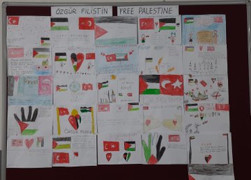 First image shows a young boy kneeling beside a small green plant in soil surrounded by a circular arrangement of child-drawn paper flags in Palestinian colors of black, white, green, and red triangles, along with heart shapes, all placed on the ground near a chain-link fence and stone border in an outdoor school area. Second image depicts several child-drawn paper hearts and Palestinian flags hanging from a chain-link fence in a rainy outdoor schoolyard with a small olive plant in the foreground and buildings in the background. Third image displays a bulletin board covered with numerous childrens crayon drawings including Palestinian flags, hearts, doves, olive branches, and text messages supporting free Palestine in Turkish and English, featuring symbols like hands and Turkish flags alongside. Fourth image captures a classroom scene with uniformed schoolchildren at desks drawing on white paper with crayons and markers, creating shapes like hearts and flags, surrounded by school supplies such as backpacks, colored pencils, and glue sticks on red-trimmed tables.