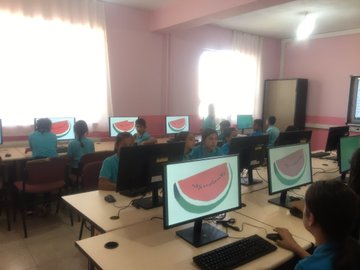 First image shows a classroom with pink walls, students in blue uniforms sitting at desks with computers displaying Palestinian flags on screens, some students working on keyboards. Second image depicts students in blue uniforms seated at desks in a pink-walled classroom, watching a smartboard showing a map or scene related to Palestine, with a teacher present. Third image features students in blue uniforms using computers with Palestinian flags on screens in a classroom with pink walls and windows. Fourth image illustrates a classroom with pink walls, students in blue uniforms at desks, a teacher in blue pointing at a smartboard displaying a hand gesture or map, engaging the class.