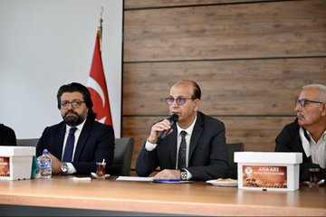 First image shows four men in suits seated at a wooden table in a meeting room with Turkish flags, holding microphones, glasses of tea, water bottles, and plates with pastries, branded boxes labeled ANA ARI on the table, wooden wall background. Second image depicts eight men in formal attire sitting around a long wooden table in a modern room with wooden walls, spotlights, and flags including Turkish and another national flag, plates and cups on the table. Third image features two men, one in white shirt and one in suit, standing on a stage holding a certificate framed with yellow border, projector screen displaying ANA ARI YETISTIRICILIGI in yellow and green, stacks of white boxes nearby on the floor. Fourth image displays a large group of about 20 diverse individuals including men and women in various attire like suits, abayas, and casual clothes, standing in a semi-circle in a spacious hall with white walls, ceiling lights, and Arabic signage, many holding certificates.