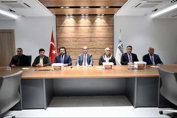 First image shows four men in suits seated at a wooden table in a meeting room with Turkish flags, holding microphones, glasses of tea, water bottles, and plates with pastries, branded boxes labeled ANA ARI on the table, wooden wall background. Second image depicts eight men in formal attire sitting around a long wooden table in a modern room with wooden walls, spotlights, and flags including Turkish and another national flag, plates and cups on the table. Third image features two men, one in white shirt and one in suit, standing on a stage holding a certificate framed with yellow border, projector screen displaying ANA ARI YETISTIRICILIGI in yellow and green, stacks of white boxes nearby on the floor. Fourth image displays a large group of about 20 diverse individuals including men and women in various attire like suits, abayas, and casual clothes, standing in a semi-circle in a spacious hall with white walls, ceiling lights, and Arabic signage, many holding certificates.