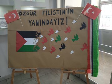 A large cardboard display stands on easels with a banner reading Ozgur Filistinin Yanindayiz in Turkish, flanked by Turkish and Palestinian flags, featuring the Palestinian flag made of paper with colorful bird cutouts in red, green, white, and black symbolizing peace. Childrens artwork covers a bulletin board including hand-drawn Palestinian and Turkish flags, houses, trees, and text in Turkish like YUNUSG K and BEREKETLI ISRAIL, with Israeli flag drawings and peace messages. Students in school uniforms sit at desks in a sunlit classroom watching a TV screen displaying Arabic text and an image of the Al-Aqsa Mosque, with bookshelves and educational posters on walls. A teacher in red stands presenting to students at desks, with a smartboard showing an image of children and Arabic text, classroom with windows and clock.