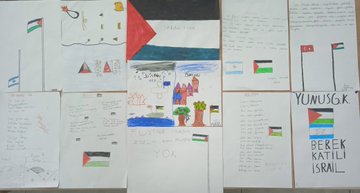 A large cardboard display stands on easels with a banner reading Ozgur Filistinin Yanindayiz in Turkish, flanked by Turkish and Palestinian flags, featuring the Palestinian flag made of paper with colorful bird cutouts in red, green, white, and black symbolizing peace. Childrens artwork covers a bulletin board including hand-drawn Palestinian and Turkish flags, houses, trees, and text in Turkish like YUNUSG K and BEREKETLI ISRAIL, with Israeli flag drawings and peace messages. Students in school uniforms sit at desks in a sunlit classroom watching a TV screen displaying Arabic text and an image of the Al-Aqsa Mosque, with bookshelves and educational posters on walls. A teacher in red stands presenting to students at desks, with a smartboard showing an image of children and Arabic text, classroom with windows and clock.