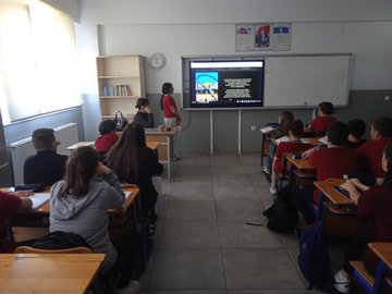 A large cardboard display stands on easels with a banner reading Ozgur Filistinin Yanindayiz in Turkish, flanked by Turkish and Palestinian flags, featuring the Palestinian flag made of paper with colorful bird cutouts in red, green, white, and black symbolizing peace. Childrens artwork covers a bulletin board including hand-drawn Palestinian and Turkish flags, houses, trees, and text in Turkish like YUNUSG K and BEREKETLI ISRAIL, with Israeli flag drawings and peace messages. Students in school uniforms sit at desks in a sunlit classroom watching a TV screen displaying Arabic text and an image of the Al-Aqsa Mosque, with bookshelves and educational posters on walls. A teacher in red stands presenting to students at desks, with a smartboard showing an image of children and Arabic text, classroom with windows and clock.