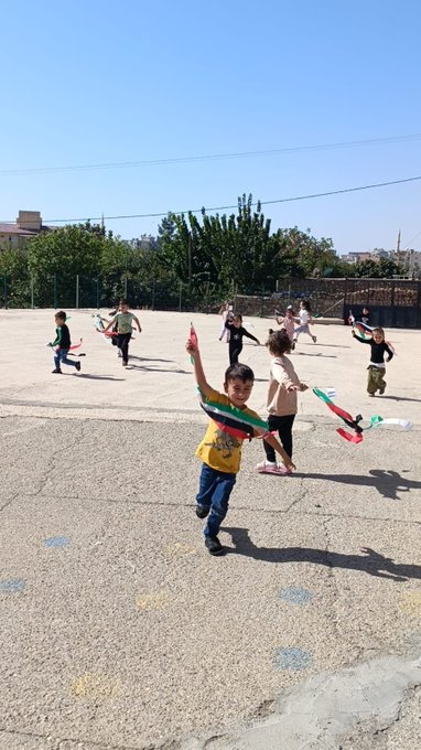 First image shows the front of a school building with a blue sign reading CUMHURIYET ILKOKULU and the Turkish flag emblem, children in colorful clothing standing on steps holding and waving Palestinian and Turkish flags. Second image depicts children on a sunny outdoor playground running and playing with Palestinian flags, some barefoot, against a background of school buildings and trees. Third image features a girl coloring a detailed drawing of Palestinian map, Dome of the Rock, ships, tents, and the number 68 on a checkered table with crayons, pencils, a water bottle, and toys nearby. Fourth image shows another girl at a table cutting or coloring a similar artwork with Palestinian symbols, ships, a girl in headscarf, flags, and landmarks on a blue mat with school bags and supplies around.