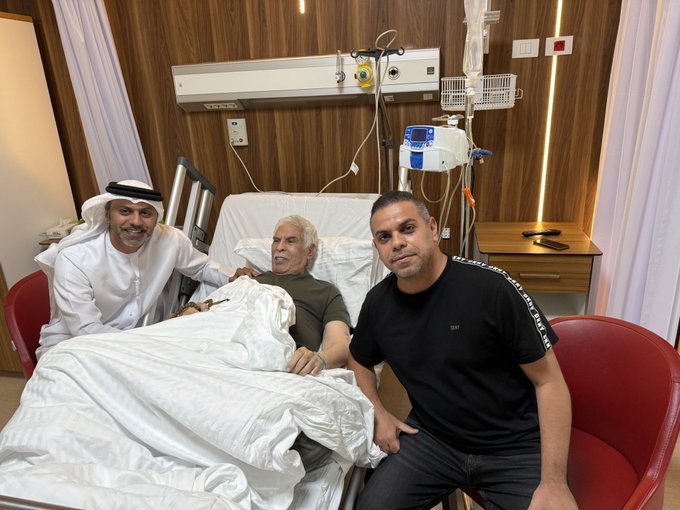 Three men in a hospital room with wooden walls and white curtains. One man in traditional white robe and headscarf stands smiling next to a hospital bed where an elderly man with white hair lies under white sheets connected to IV drip and medical equipment. Another man in black t-shirt and pants sits on a red chair beside the bed smiling. Medical monitors and tubes are visible on the bedside table.