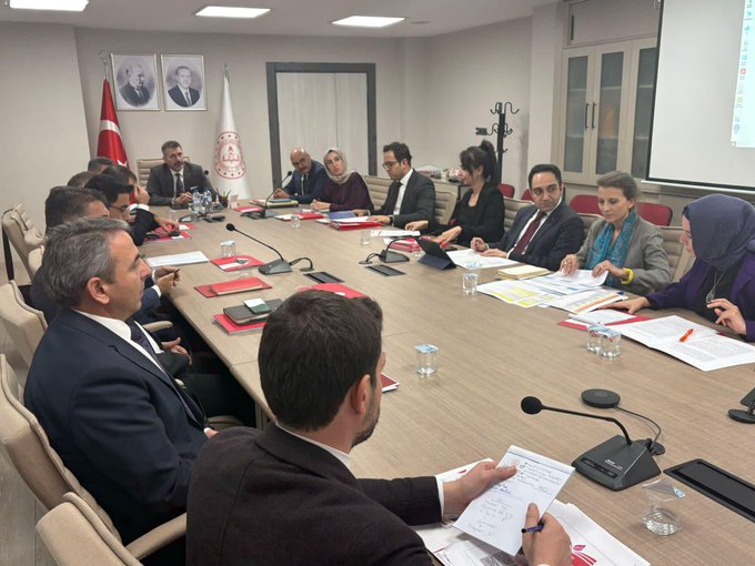 İlk görüntüde resmi bir toplantı odasında uzun bir masa etrafında oturan yaklaşık 12-15 kişi görülüyor, hepsi iş kıyafetleri giymiş erkekler takım elbiseli kadınlar başörtülü bazıları, masada mikrofonlar kırmızı dosyalar su bardakları ve Türk bayrağı ile başka bir bayrak duvarda asılı, arka planda iki portre asılı ve pencere perdeleri var. İkinci görüntüde benzer bir toplantı odasında masa etrafında 10-12 kişi oturuyor, mikrofonlar belgeler su bardakları kırmızı dosyalar mevcut, duvarda Türk bayrağı ve başka bayraklar portreler asılı, bir projektör ekranı boş görünüyor ve bazı kişiler not alıyor.