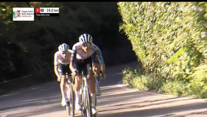 Two cyclists in white and pink team jerseys and helmets ride closely on a tree-lined road during a race. The rider in front wears number 1 on his back. Race graphics overlay shows Giro dItalia logo, stage 10, 230 km distance, and 28 km remaining. Green foliage borders the asphalt path under daylight.