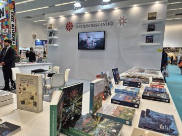 İlk resimde çeşitli katılımcılar İletişim Başkanlığı standı önünde poz veriyor kitaplar masada dizili Türk bayrağı ekranda görünüyor. İkinci resimde grup halinde resmi kıyafetli kişiler stand önünde kitaplar ve Türkiye Cumhuriyeti Cumhurbaşkanlığı İletişim Başkanlığı yazısı masada. Üçüncü resimde stand kitaplarla dolu YÜKSEK standı arka planda bir adam kitaplara bakıyor. Dördüncü resimde boş stand kitaplar masada sandalye ve ekran fuar ortamı.