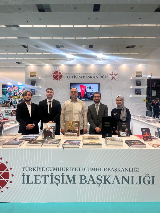 İlk resimde çeşitli katılımcılar İletişim Başkanlığı standı önünde poz veriyor kitaplar masada dizili Türk bayrağı ekranda görünüyor. İkinci resimde grup halinde resmi kıyafetli kişiler stand önünde kitaplar ve Türkiye Cumhuriyeti Cumhurbaşkanlığı İletişim Başkanlığı yazısı masada. Üçüncü resimde stand kitaplarla dolu YÜKSEK standı arka planda bir adam kitaplara bakıyor. Dördüncü resimde boş stand kitaplar masada sandalye ve ekran fuar ortamı.