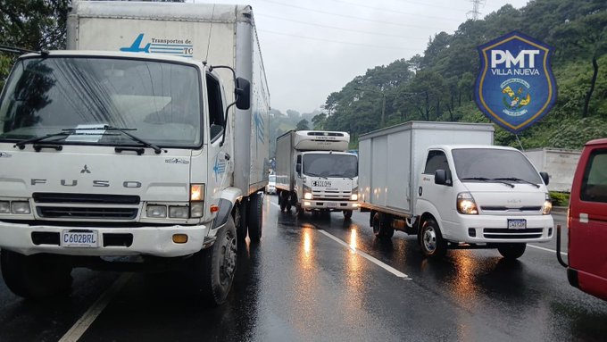 Multiple white and silver trucks including a large Fuso truck with license plate visible are lined up on a wet two-lane highway under rainy conditions with water on the road surface. A small white van and another truck are in the background. A blue shield emblem labeled PNMT VILLA NUEVA is displayed on a pole beside the road. Trees and hills are visible in the misty background.
