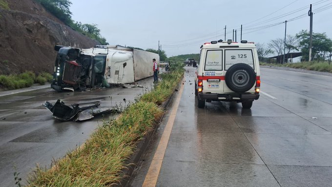 Overturned white trailer truck lies on its side on a wet roadside with grass and a dirt embankment nearby. Red and white Cruz Roja ambulance van with license plate visible parks adjacent to the wreckage. Debris scatters across the pavement under rainy conditions. Responders in uniforms stand near the scene. Second image shows similar setup with a person walking toward the truck from the ambulance side. Gray sedan parked nearby. Roadway lined with utility poles and vegetation.