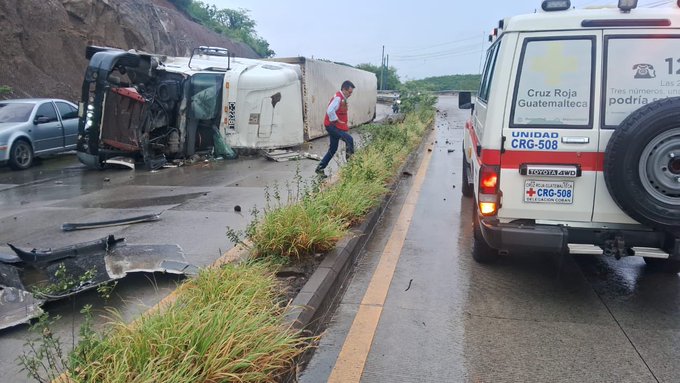 Overturned white trailer truck lies on its side on a wet roadside with grass and a dirt embankment nearby. Red and white Cruz Roja ambulance van with license plate visible parks adjacent to the wreckage. Debris scatters across the pavement under rainy conditions. Responders in uniforms stand near the scene. Second image shows similar setup with a person walking toward the truck from the ambulance side. Gray sedan parked nearby. Roadway lined with utility poles and vegetation.